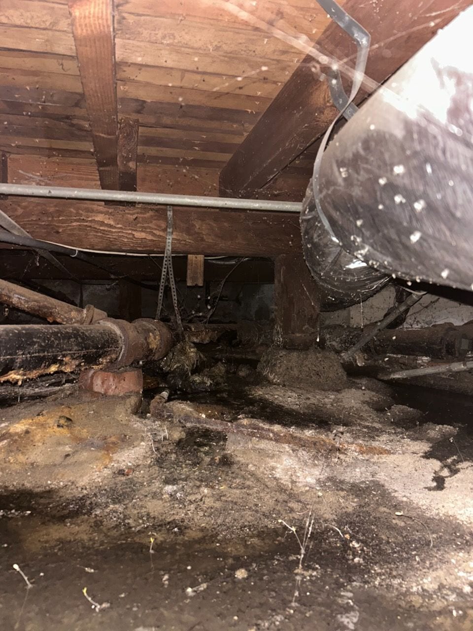 Wet, debris-covered crawlspace under a home with ductwork and old plumbing overhead.
