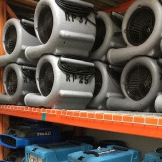 Restoration Pros air movers and dehumidifiers stacked on warehouse shelving.