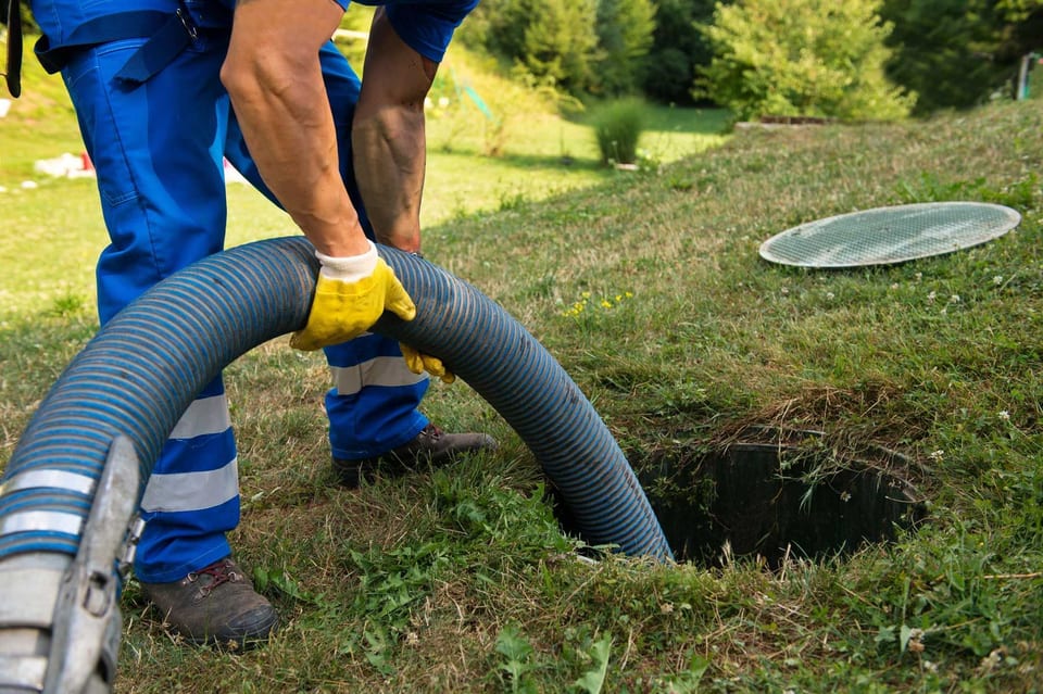 Worker feeding a large vacuum hose into an underground tank opening in a yard.