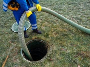 Worker in coveralls feeding a vacuum hose into an open sewer access on a lawn.